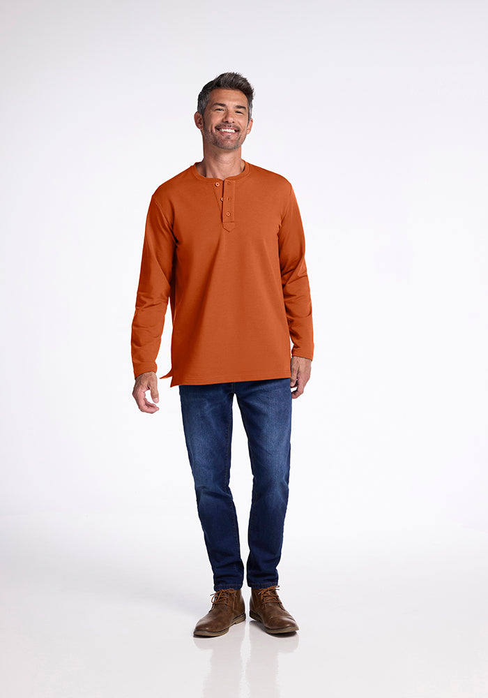 A smiling man with short, dark hair is wearing a Woolx Jackson Henley in burnt orange, paired with dark blue jeans and brown shoes. He stands against a plain white background, looking relaxed with his arms by his sides.