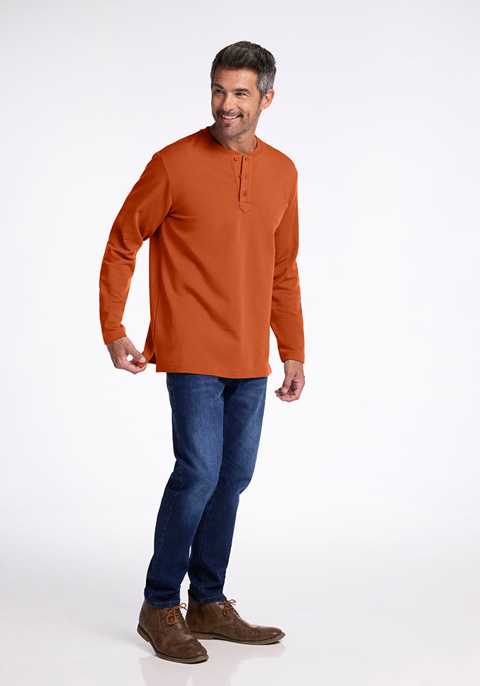 A man stands facing slightly right, smiling, wearing a long-sleeved Jackson Henley in orange by Woolx, blue jeans, and brown shoes. The background is a plain white studio backdrop.