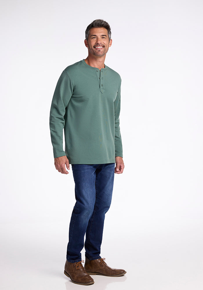 A man with short gray hair and a beard is smiling and standing. He is wearing the Jackson Henley in Duck Green by Woolx, along with dark blue jeans and brown shoes. The background is a plain white studio setting.