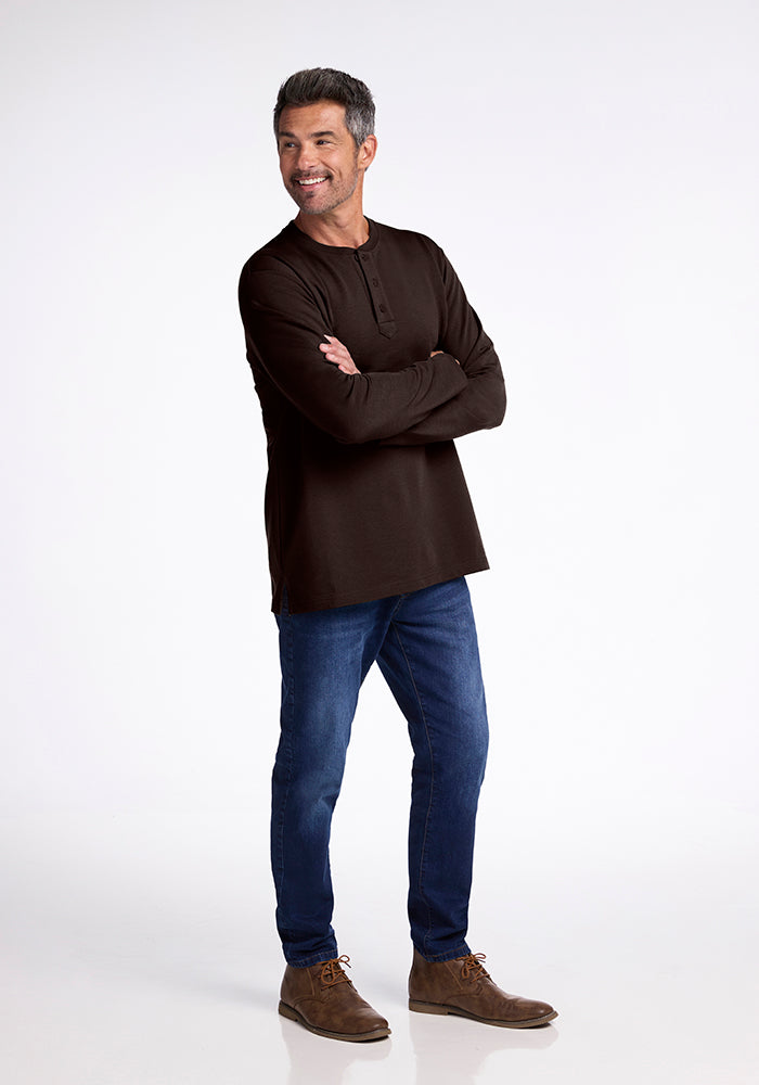 A man wearing a black Jackson Henley by Woolx and blue jeans stands with arms crossed, smiling and looking to the side. He is sporting brown shoes and has short, gray hair, set against a plain white background.