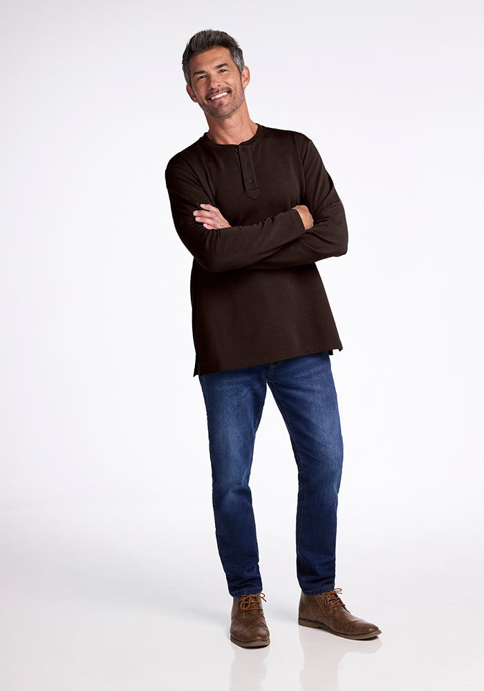 A man stands with a relaxed posture, dressed in a dark Jackson Henley shirt from Woolx and blue jeans. He has short, dark hair with gray streaks and is smiling slightly. He is wearing brown lace-up shoes. The background is plain white.