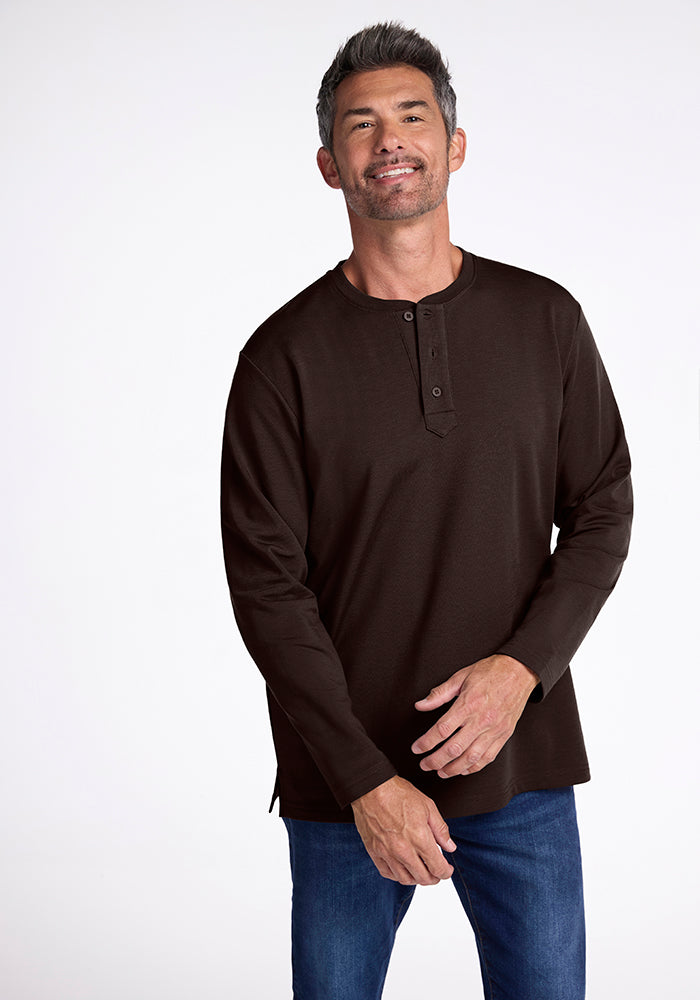 A man with short, graying hair and a beard is wearing a long-sleeved Jackson Henley in dark brown, made by Woolx, paired with blue jeans. He is standing against a plain white background, smiling slightly and looking directly at the camera. 