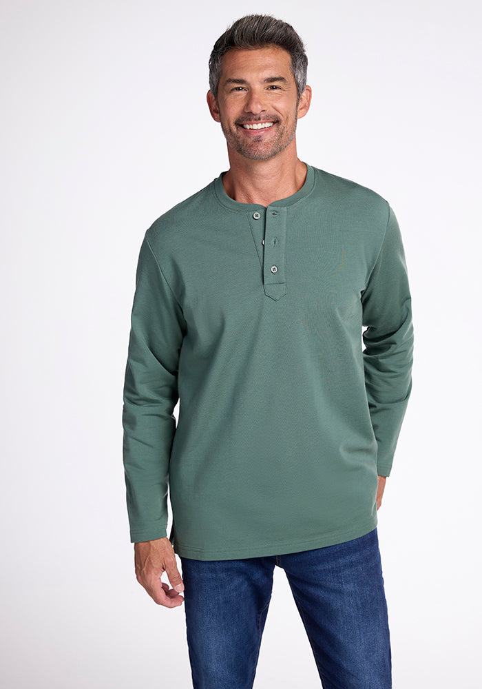 A person with short gray hair and a slight beard smiles while wearing a long-sleeved green Jackson Henley by Woolx and blue jeans. They are standing against a plain white background. 