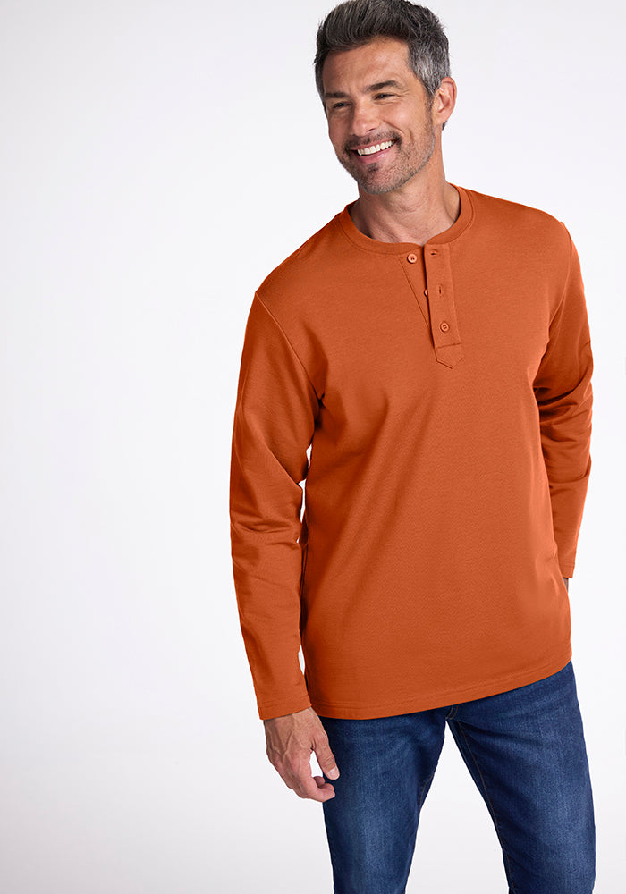 A man with short, graying hair smiles while wearing an orange Jackson Henley by Woolx. He is also wearing blue jeans. The background is plain white. 