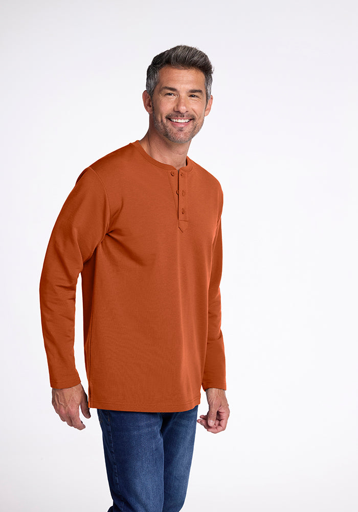 A man with short, dark hair and a beard is smiling at the camera. He is wearing an orange long-sleeved Jackson Henley by Woolx made of midweight merino wool and blue jeans. The background is plain and light-colored.