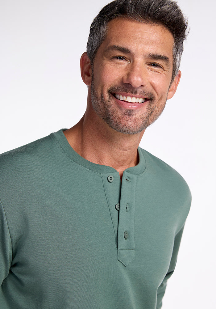 A man with short gray hair and a beard is smiling at the camera. He is wearing the Jackson Henley in Duck Green from Woolx, made from Australian Merino Wool, with three buttons undone. The background is plain white.