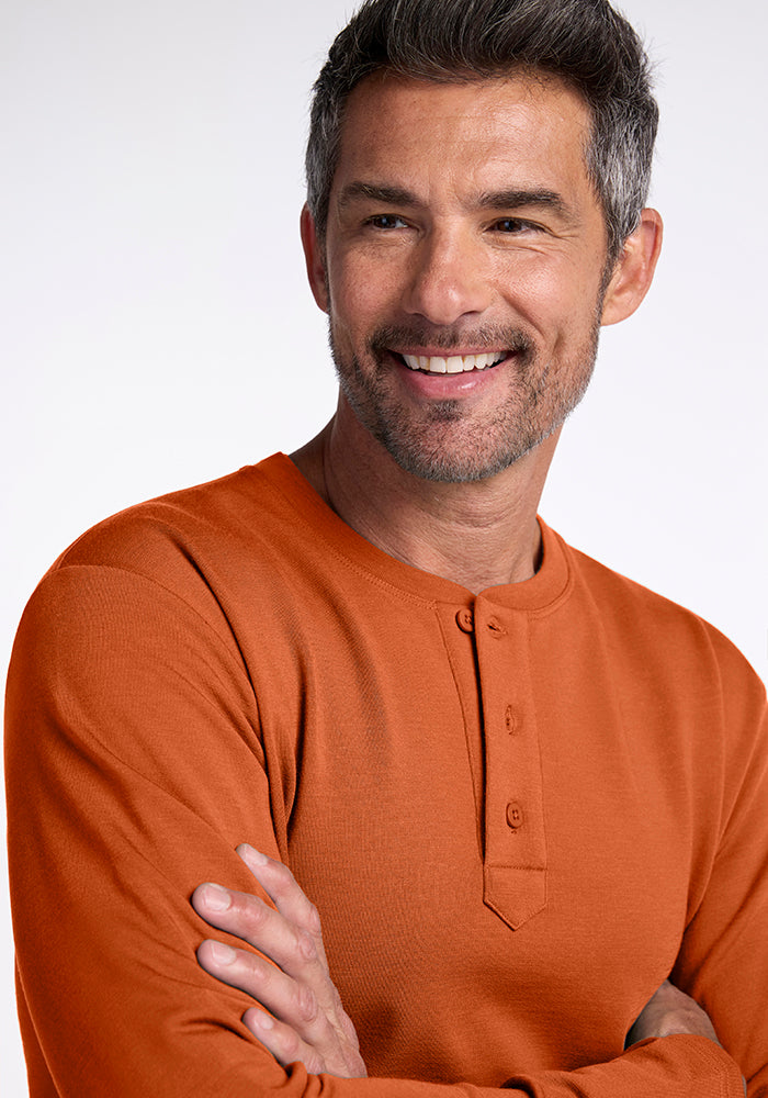 A man with short graying hair and a beard smiles and looks off to the side. He is wearing a long-sleeved Jackson Henley in orange, made from midweight merino wool by Woolx, and stands with his arms crossed against a light background.