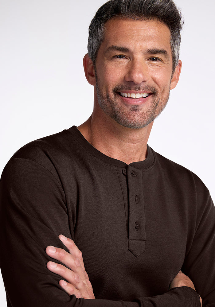 A man with short, salt-and-pepper hair and a beard is smiling with his arms crossed. He is wearing the Jackson Henley, a dark brown mid/heavyweight Merino wool shirt with a buttoned placket by Woolx. The background is plain and light-colored.