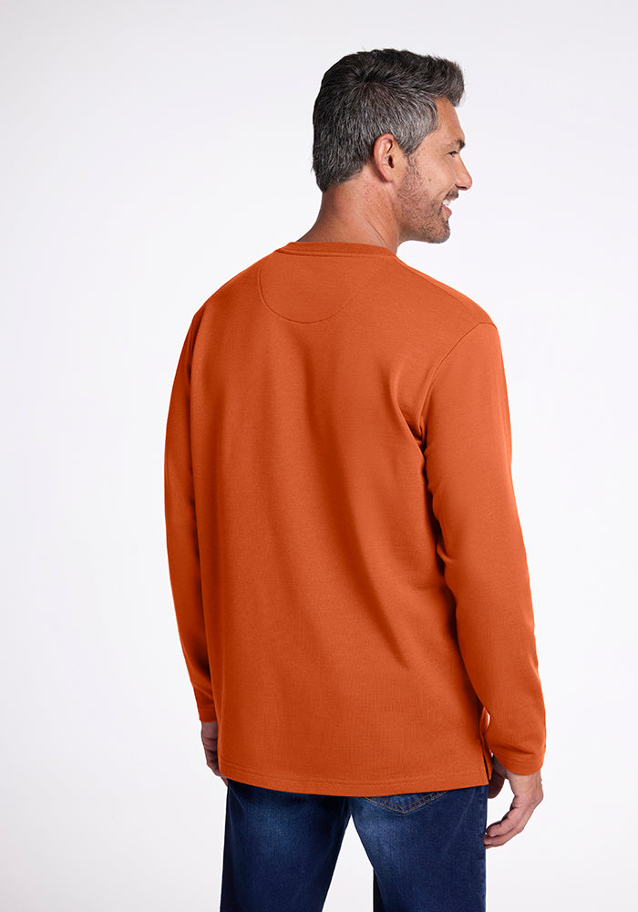 A man with short gray hair is standing and facing away, looking to the right. He is wearing an orange Jackson Henley shirt from Woolx, made of mid/heavyweight Merino wool, paired with dark blue jeans. The background is plain white.