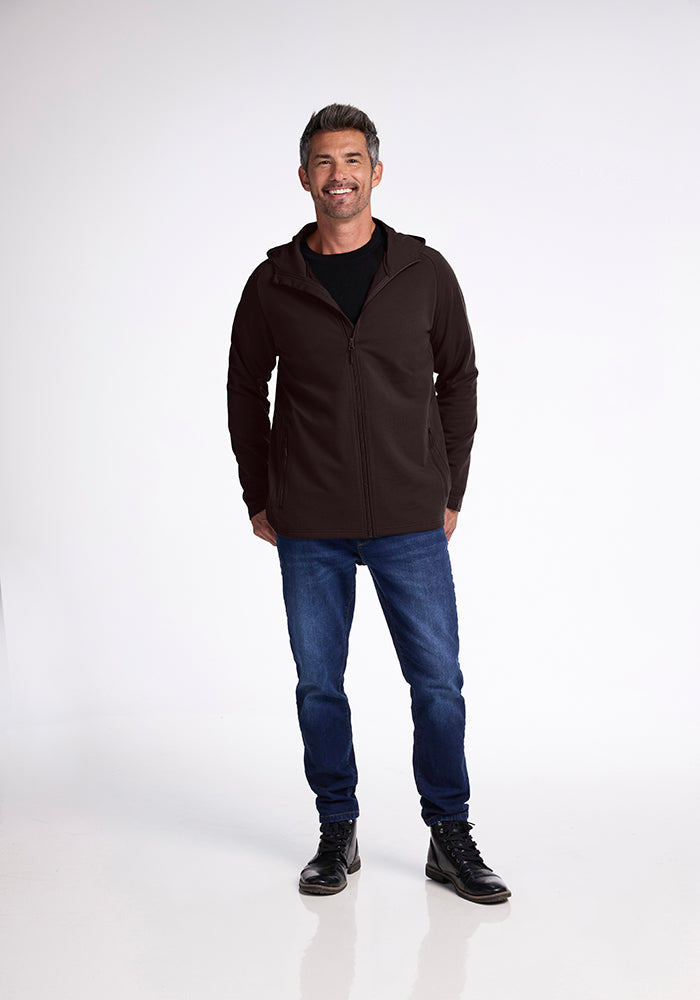 A man smiling, standing with hands in his pockets, wearing a French Roast Boulder Hoodie from Woolx, dark blue jeans, and black boots. He has short, dark hair and is standing in front of a plain white background.