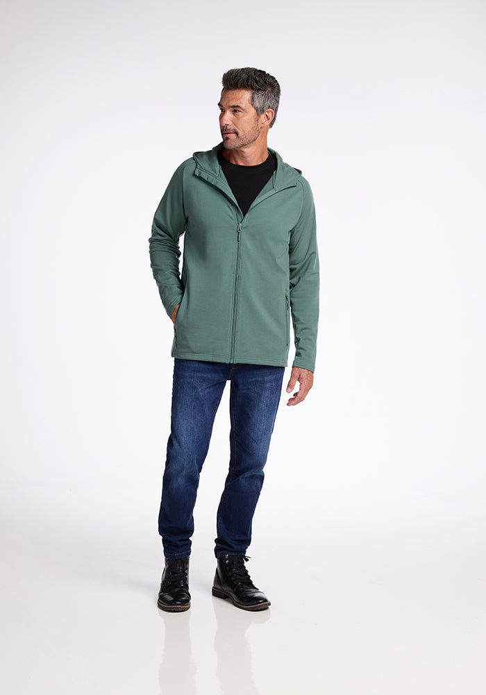 A man stands in a studio setting, wearing the Duck Green Boulder Hoodie from Woolx over a black shirt, blue jeans, and black boots. He is looking off to the side with his left hand in his pocket. The background is plain and white.