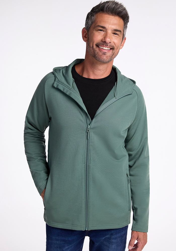 A smiling man with gray hair is wearing a Duck Green, breathable Merino wool Boulder Hoodie from Woolx over a black t-shirt. He has his left hand in his hoodie pocket. The background is plain and white.