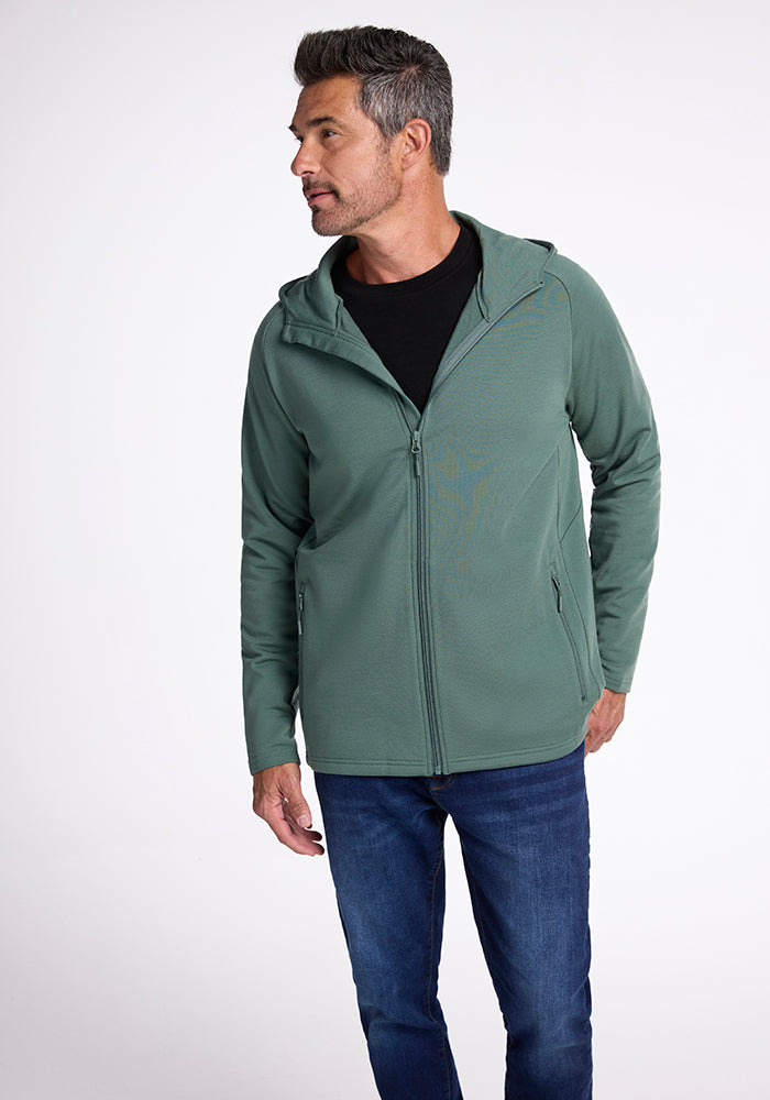 A man stands facing slightly to the left, wearing a Woolx Boulder Hoodie in Duck Green over a black shirt and blue jeans. He has short, greying hair and a goatee, and is looking off to the side with a neutral expression.