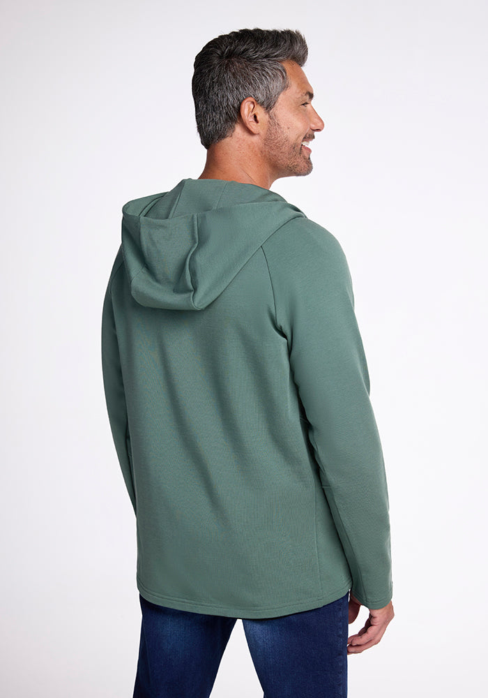 A man with short gray hair smiles as he looks to his left, wearing the Boulder Hoodie - Duck Green from Woolx, made of breathable fabric, and blue jeans. The background is plain white. He is positioned with his back turned slightly to the camera, showcasing the back and side of the hoodie.