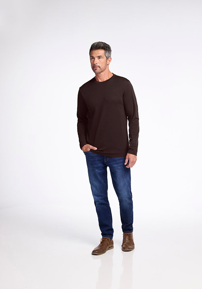 A man with short gray hair stands against a plain white background, wearing a Woolx Easton Shirt, blue jeans, and brown shoes. He looks off to the side with a contemplative expression.