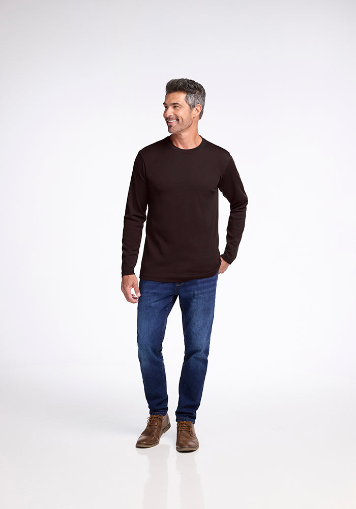 A man with short gray hair, wearing the Woolx Easton long-sleeved black midweight shirt made from Australian Merino Wool, blue jeans, and brown shoes, stands smiling against a plain white background. He is relaxed with one hand in his pocket and the other hanging loosely by his side.