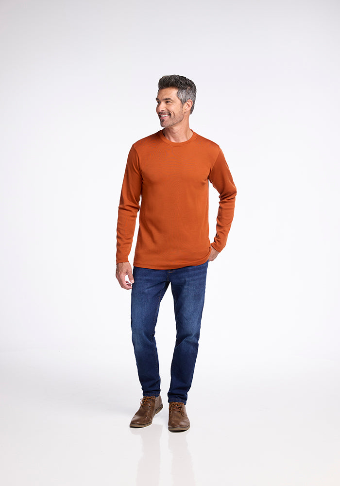 A man with short hair is standing and smiling, wearing the Easton Shirt in Cinnamon Stick from Woolx, along with dark blue jeans and brown shoes. He is facing slightly to the right and has his left hand in his pocket against a plain white background.