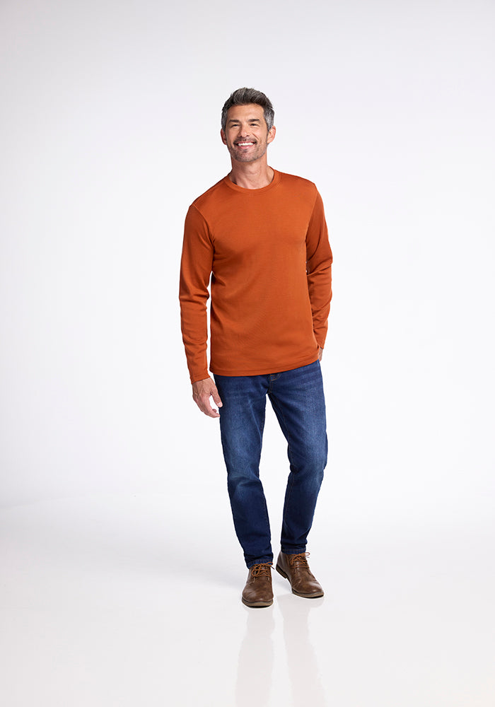 A man with short hair is standing against a plain white background, smiling slightly. He is wearing Woolx's long-sleeved, midweight 230g/m² Easton Shirt in orange, blue jeans, and brown leather shoes. His hands are relaxed by his sides.