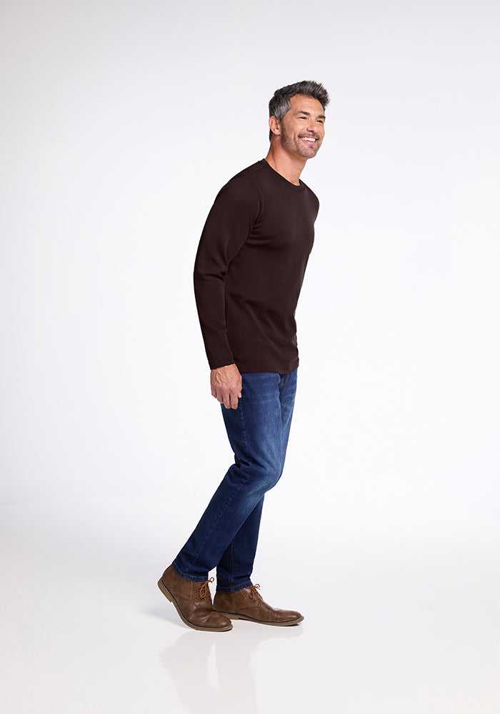 A man stands against a plain white background, smiling. He is wearing the Easton Shirt from Woolx, blue jeans, and brown shoes. He leans slightly forward, balancing on his right leg with his left leg slightly lifted behind him. His hands are by his sides.