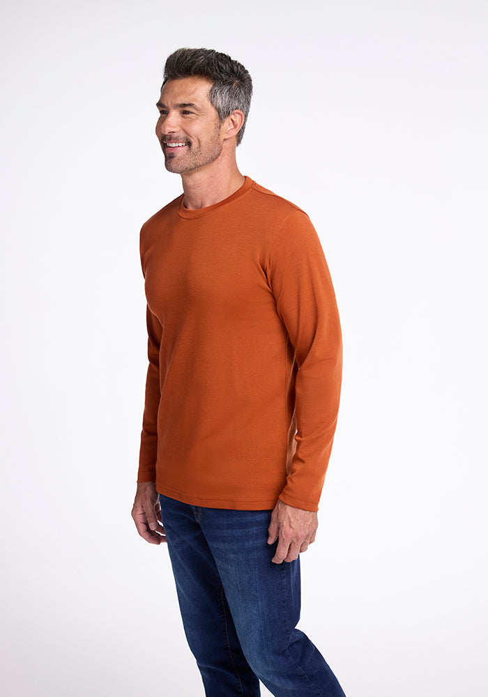 A person with short, graying hair and a trimmed beard is wearing Woolx's Easton Shirt, a long-sleeved, orange shirt made from midweight 230g/m² Australian Merino Wool, along with blue jeans. They are standing and smiling, facing slightly to the left against a plain, white background.