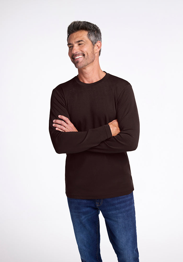 A man with short gray hair and a beard is smiling and looking to the side. He is wearing a long-sleeve, black Easton Shirt by Woolx, made from Australian Merino Wool, paired with blue jeans. His arms are crossed over his chest, and the background is plain white.