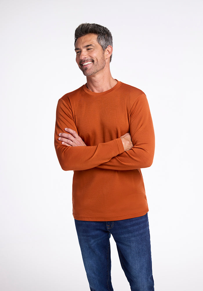 A man with short salt and pepper hair and a beard is standing with his arms crossed, smiling. He is wearing an orange long-sleeve Easton Shirt by Woolx and blue jeans made from Australian Merino Wool. The background is plain white. 