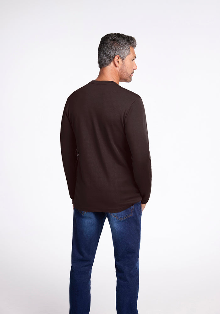 A man with short, gray hair stands facing away from the camera. He is wearing a long-sleeve, dark brown Easton Shirt in French Roast by Woolx and blue jeans. The background is plain and light-colored, creating a stark contrast with his attire.