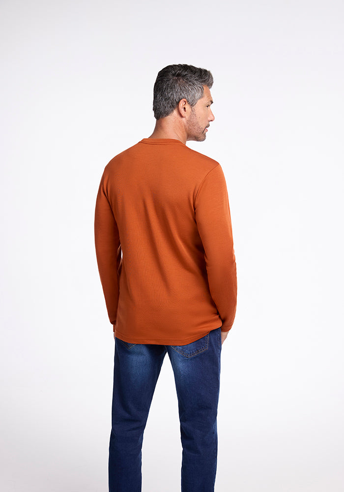 A man with short, gray hair stands with his back to the camera, wearing a Woolx Easton Shirt in Cinnamon Stick and blue jeans. The background is a plain, light gray color. Beneath his clothes, he sports a Merino base layer for added warmth.