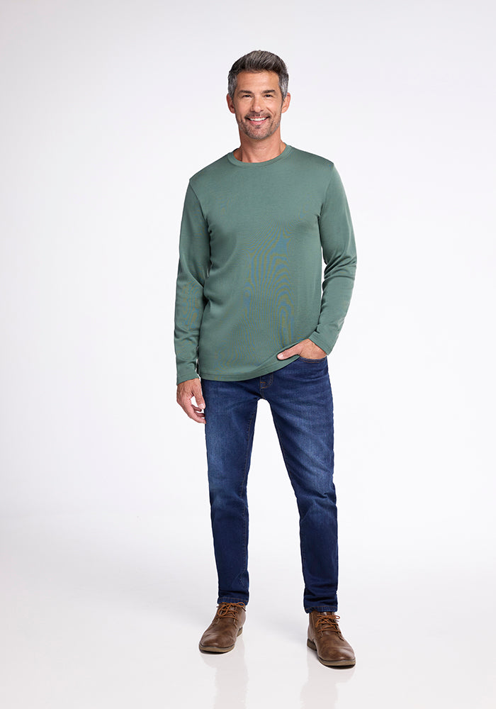 A man stands against a white background, smiling. He has short gray hair and is wearing the Woolx Easton Shirt in Duck Green, dark blue jeans, and brown shoes. With one hand in his jeans pocket, he appears relaxed, exuding comfort and exceptional moisture management.