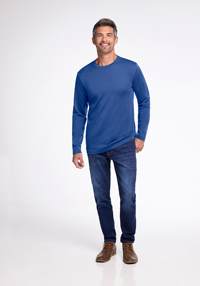 A man with gray hair and a trimmed beard is smiling as he poses. He is wearing a blue Easton Shirt by Woolx, made of Australian Merino Wool, along with dark blue jeans and brown shoes. The background is a plain, light-colored backdrop.