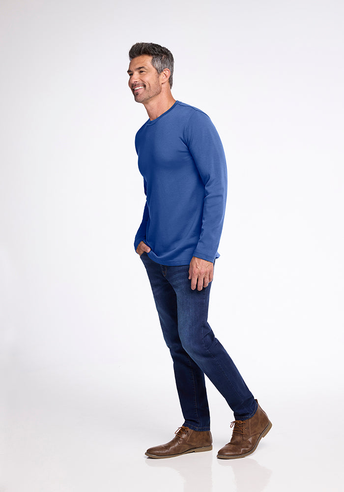 A man with short, gray hair is posing in a relaxed stance. He is wearing a long-sleeved, blue Easton Shirt from Woolx made from Australian Merino Wool, dark jeans, and brown lace-up shoes. The background is plain white.
