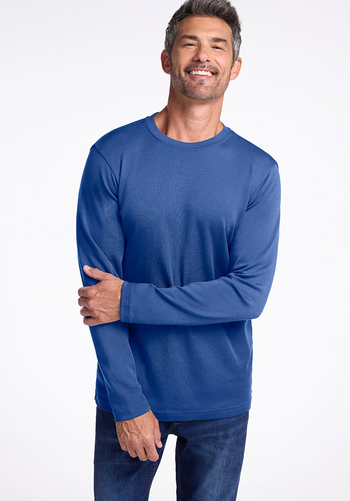 A man with short gray hair and a beard smiles while standing against a plain white background. He is wearing the long-sleeve, blue Easton Shirt from Woolx, made from Australian Merino Wool, paired with dark jeans. His right hand rests on his left forearm.