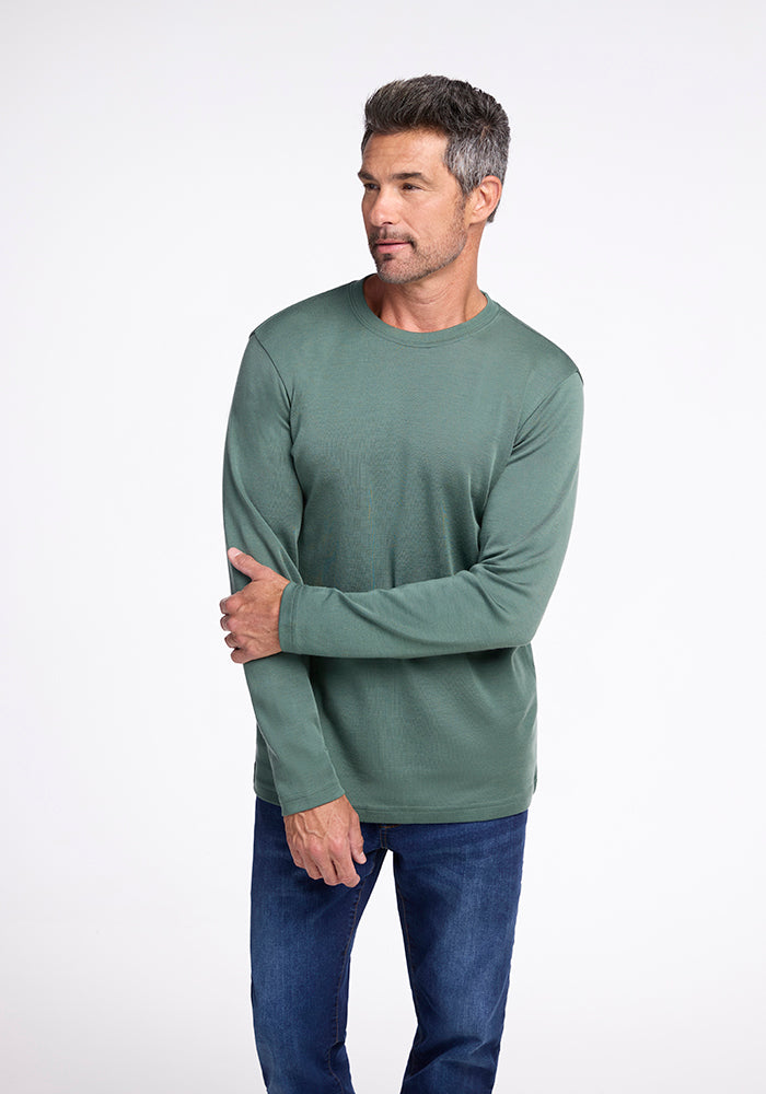 A man with short, graying hair and a trimmed beard is wearing a long-sleeved, green Woolx Easton Shirt and blue jeans. He stands against a plain white background, looking slightly to his left, with his left arm crossing his body to touch his right elbow.