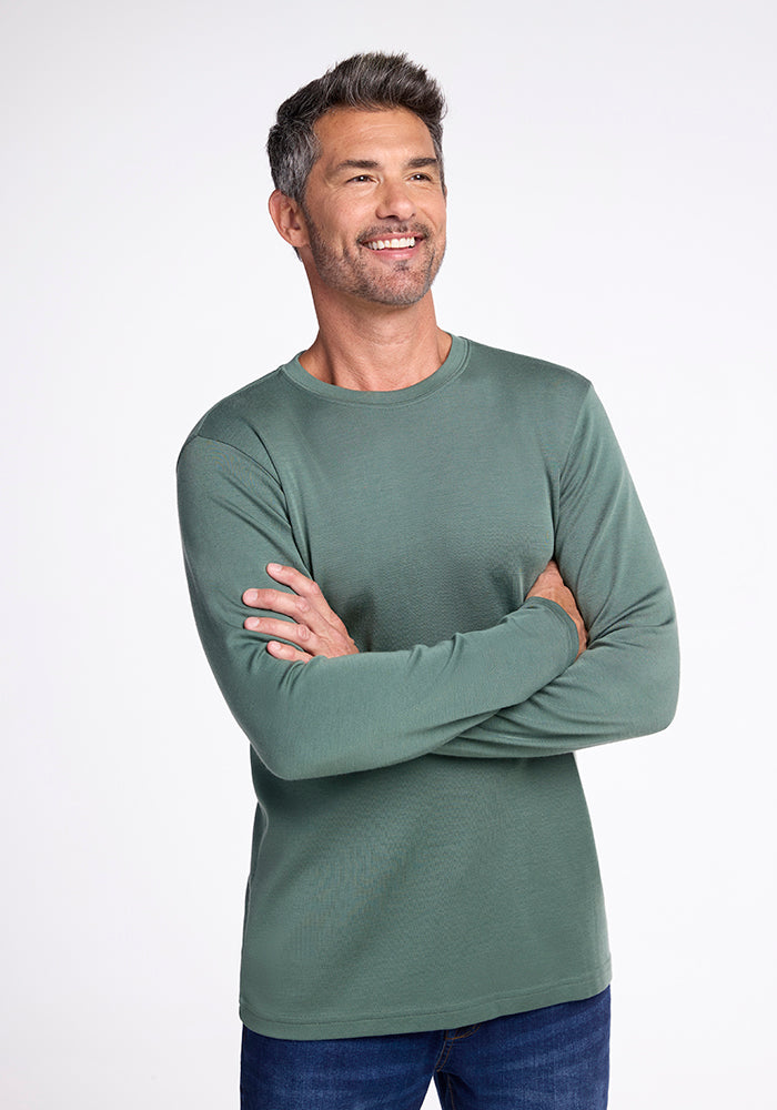 A man with salt-and-pepper hair is smiling and standing with his arms crossed. He is wearing a green long-sleeved Easton Shirt by Woolx, made from Merino wool known for its excellent moisture management, and blue jeans. The background is plain white. 