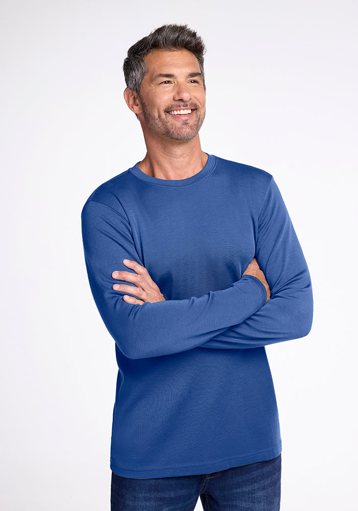 A man with short gray hair is smiling and looking slightly to the side. He is wearing the long-sleeved, blue Easton Shirt by Woolx and blue jeans. His arms are crossed over his chest, and he stands against a plain, light-colored background. 