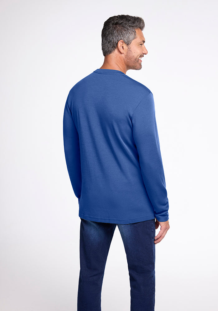 A man with short, gray hair is standing and facing away from the camera. He is wearing a long-sleeved Easton Shirt by Woolx, made of Australian Merino Wool, along with navy blue pants. The background is a plain, light-colored studio setting.