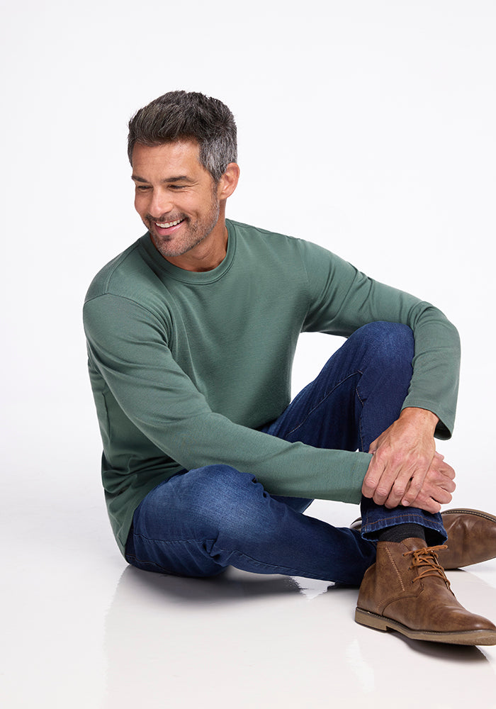 A man with short gray hair and a beard is sitting on the floor against a white background. He is wearing Woolx's green Easton Shirt, blue jeans, and brown suede boots. The shirt, made from Australian Merino Wool, showcases his effortless style. Smiling and looking to the side, he rests one hand on his knee.