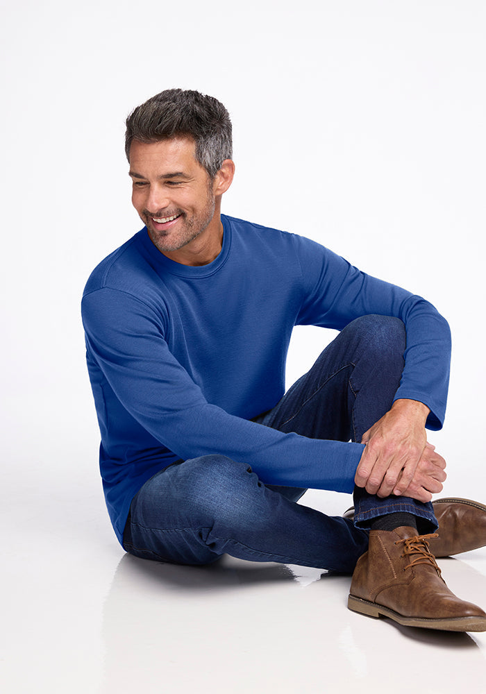 A man with short gray hair and a beard is smiling while sitting on the floor with one leg crossed over the knee. He is wearing a blue long-sleeve Easton Shirt in Oceanside color from Woolx, along with blue jeans and brown boots. The background is plain white.