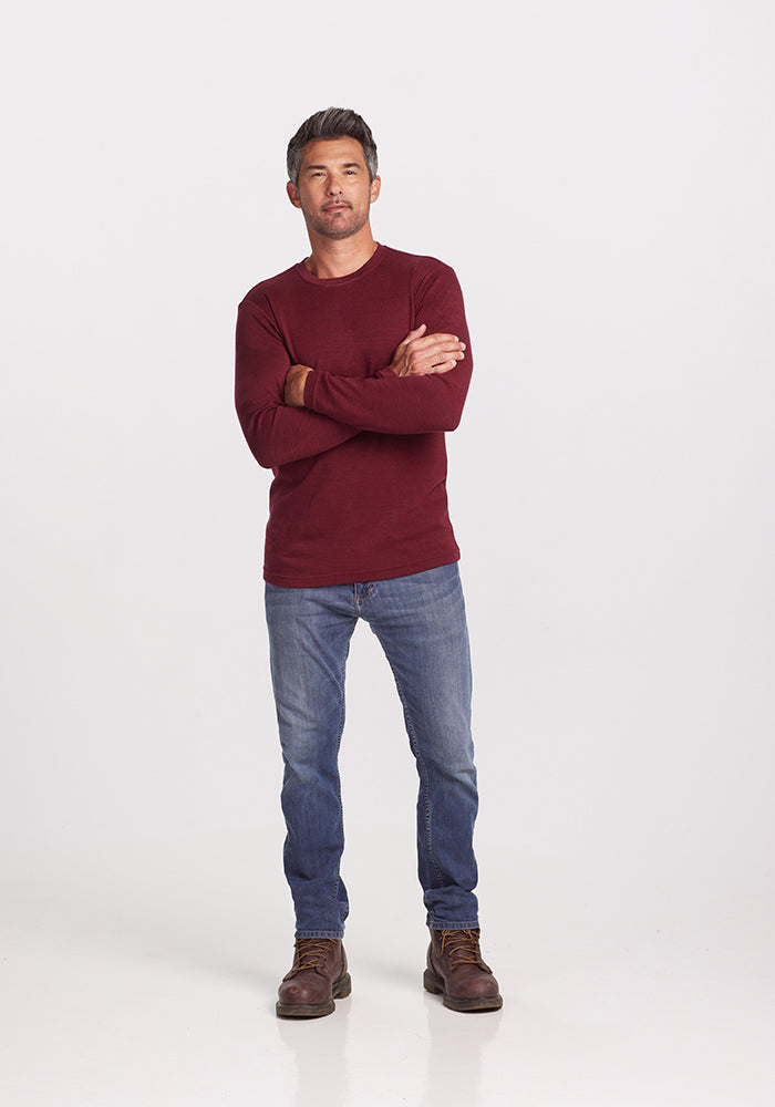 A man stands with arms crossed, wearing an Easton Shirt by Woolx, made from Australian Merino Wool. He pairs it with blue jeans and brown boots. With short dark hair and a neutral expression, he is set against a plain white background, exuding a casual style.