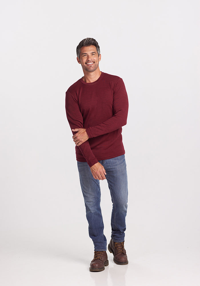 A person wearing a cranberry melange long sleeve shirt and blue jeans stands against a plain white background, smiling, with one arm relaxed across their body and the other hand holding their elbow. The cranberry shirt is the Easton Shirt by Woolx, an Australian Merino Wool base layer, paired perfectly with brown shoes for everyday wear.