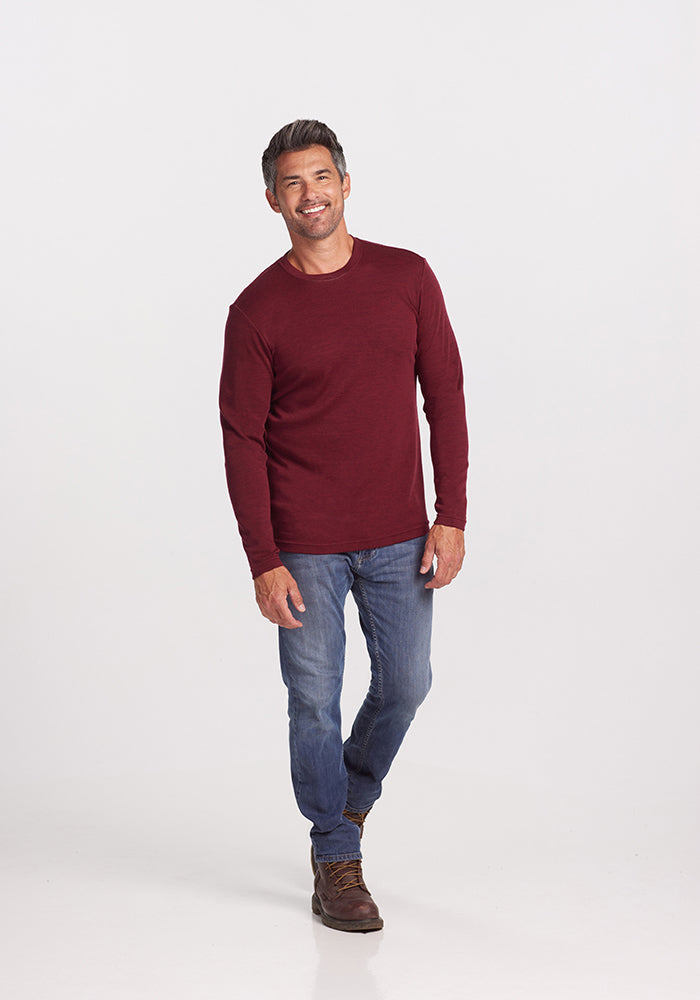 A man with short dark hair is smiling as he stands against a plain white background. He is wearing a maroon Woolx Easton Shirt made of Australian Merino Wool, blue jeans, and brown boots. Casually posing, his left hand relaxed by his side and his right hand loosely clasping his left wrist.