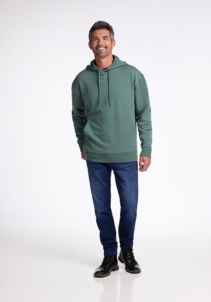 A smiling man stands on a plain white background, dressed in a cozy Duck Green Chase Hoodie by Woolx, dark blue jeans, and black shoes. His gray hair and beard add to his distinguished appearance, and his hands rest comfortably by his sides.