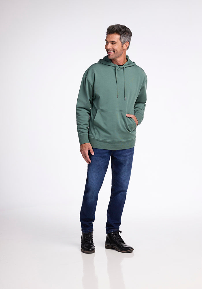 A man stands on a white background, smiling while looking to his left. He is wearing the Woolx Chase Hoodie in Duck Green, a men's hoodie with a front pocket crafted from soft merino wool. He pairs it with dark blue jeans and black boots. His left hand is casually placed in his hoodie's pocket, ready for an outdoor adventure.