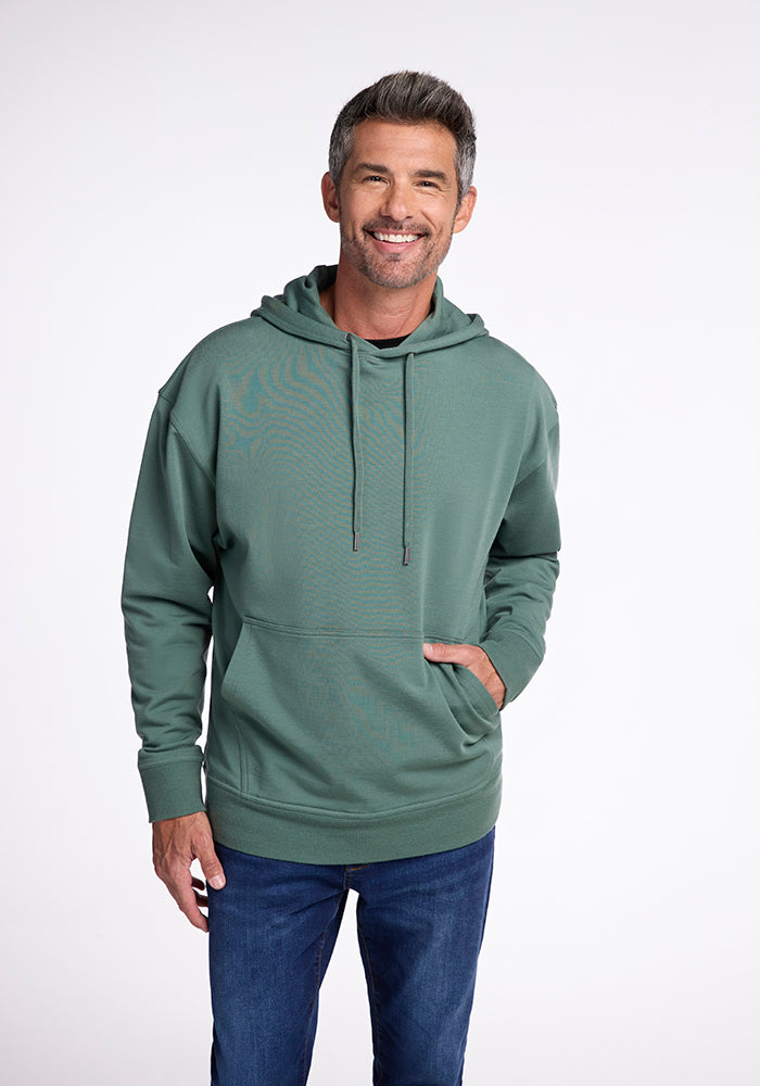 A man with short, salt-and-pepper hair, wearing a Woolx Chase Hoodie in Duck Green made of soft Merino wool, along with blue jeans, smiles with his hand in his hoodie pocket. The background is plain white.