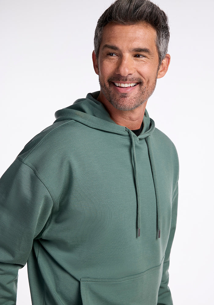 A man with short, neatly styled gray hair and a beard is smiling and looking slightly to the side. He is wearing a Duck Green Chase Hoodie made from Merino wool by Woolx. The background is plain white.