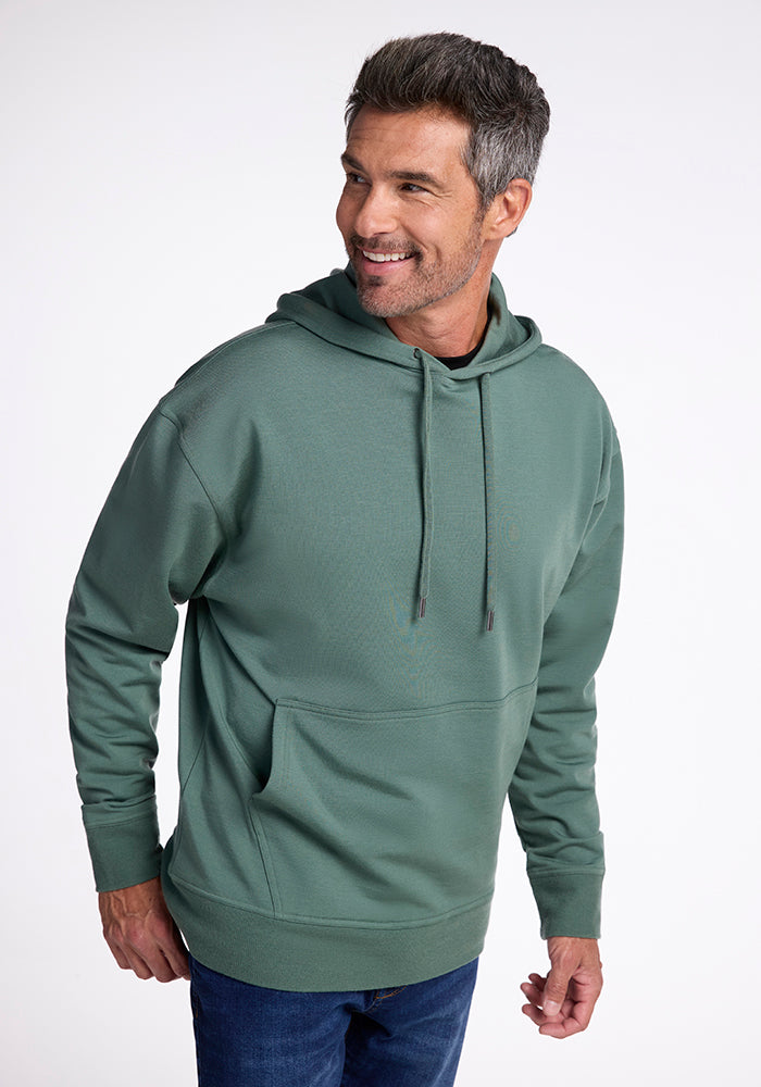 A man with short, gray hair is smiling while wearing the Chase Hoodie - Duck Green from Woolx and jeans. The adventure sweatshirt features drawstrings and a front pocket. He is standing against a plain, white background and looking slightly to his right. 