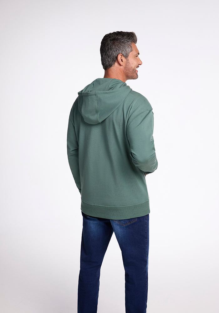 A person with short gray hair stands facing slightly left, wearing a Woolx Chase Hoodie in Duck Green, made of Merino wool, and blue jeans. The background is plain white.