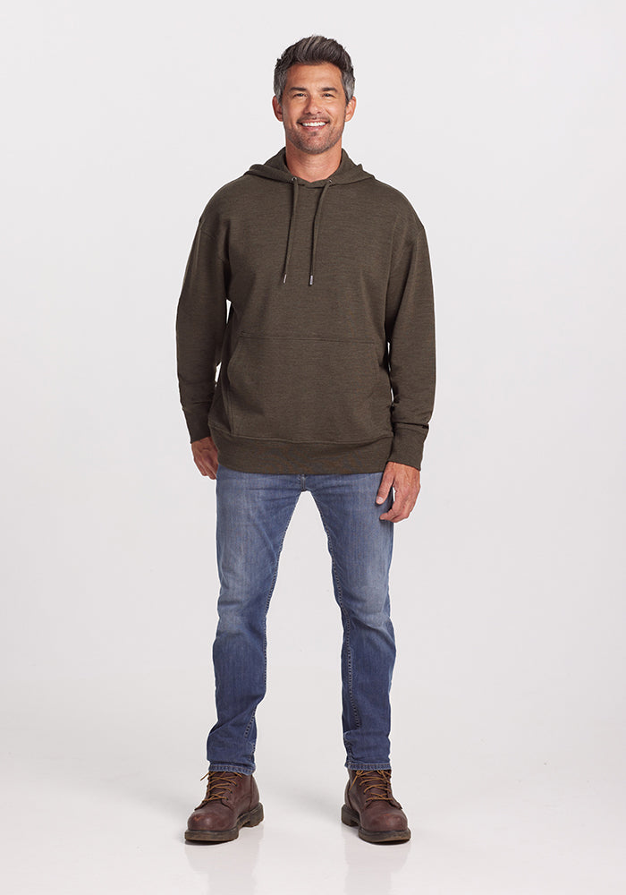 A man with short dark hair is standing and smiling. He is wearing a Woolx Chase Hoodie in Dark Moss made from odor-resistant Merino Wool, light blue jeans, and brown boots. The background is plain white.