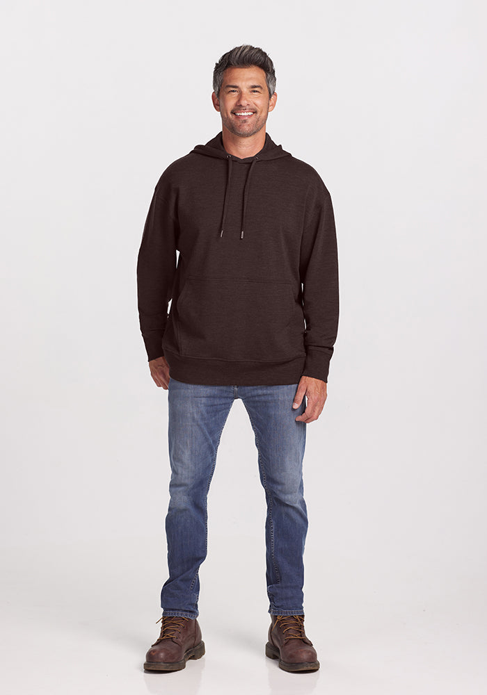 A person stands facing the camera wearing a dark Woolx Chase Hoodie in Maple Melange, blue jeans, and brown boots. They are smiling with their hands relaxed by their sides against a plain white background.