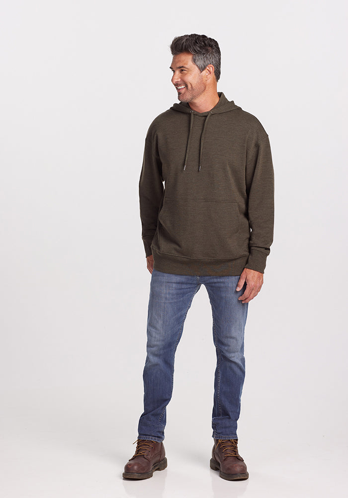 A man with short, gray hair is standing and smiling. He is wearing a dark moss, odor-resistant Merino wool Chase Hoodie by Woolx with drawstrings, blue jeans, and brown boots. The background is plain and white.